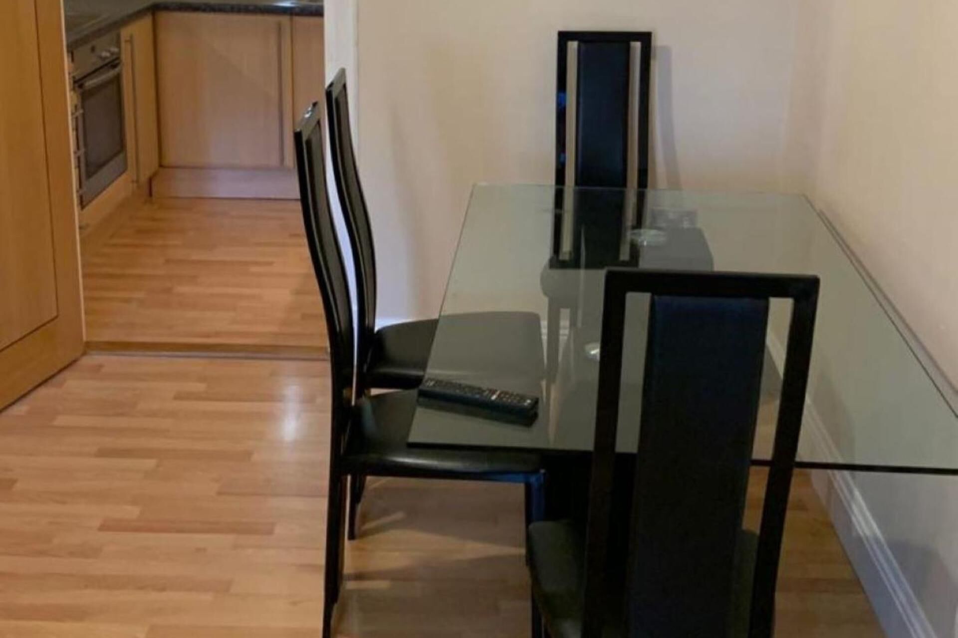 A dining area with a glass table and black chairs, a remote control on the table, and a kitchen in the background.