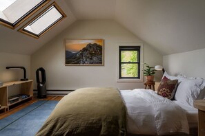 A bedroom with a skylight, a bed, a nightstand, a lamp, a potted plant, and a painting of a mountain.