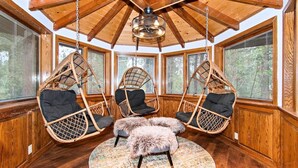 A cozy room with a high ceiling, wooden beams, and large windows. There are three wicker chairs with cushions and a fluffy rug on the floor.