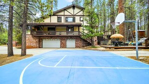 A basketball court in front of a two-story house with a balcony, garage, and outdoor seating area.