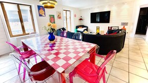 A modern living room with a checkered table, clear plastic chairs, a sofa, and a flat-screen TV. - Family & Fun! Pedestrian center near Lake and Ski (Gérardmer)