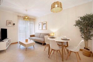 A modern living room with a sofa, dining table, chairs, a TV, and a potted plant. - Apartment 'Quinta Da Gomeira 67' with Shared Pool, Wi-Fi and Air Conditioning (Tavira)
