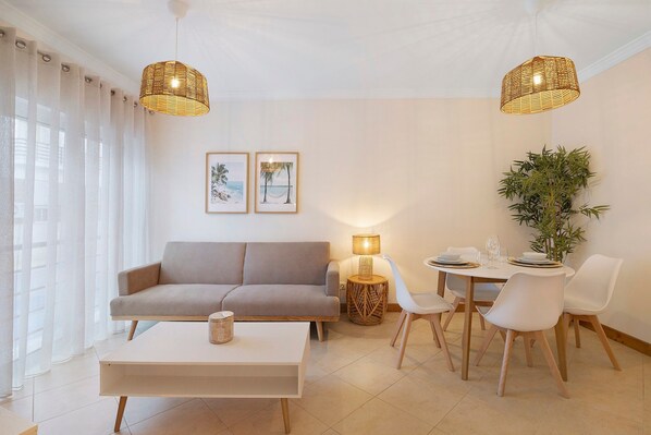 A living room with a sofa, coffee table, dining table with chairs, two hanging lights, a lamp, and framed pictures on the wall.