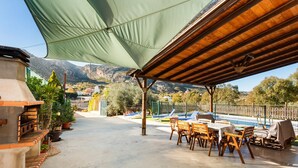 Patio esterno con tavolo e sedie, un barbecue e una zona piscina sullo sfondo.
