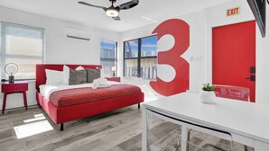 Camera d'albergo moderna con un grande numero rosso '3' sulla parete, un letto con lenzuola rosse, una scrivania e una ventola a soffitto.