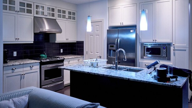 Cocina moderna con gabinetes blancos, electrodomésticos de acero inoxidable, una isla central con pileta e iluminación colgante.