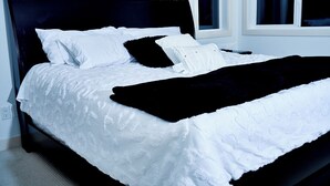 Una cama bien tendida con ropa blanca y una manta negra, ubicada en una habitación con paredes blancas y una ventana.