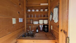A compact wooden cabin with a sink, shelves with cups and bowls, a coffee maker, and a bottle of hand soap. - Hill Country Retreat (Austin)