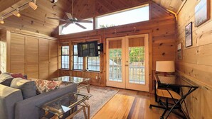 A cozy cabin interior with a wooden ceiling, a ceiling fan, a glass coffee table, a sofa, a TV mounted on the wall, and a door leading outside. - Hill Country Retreat (Austin)