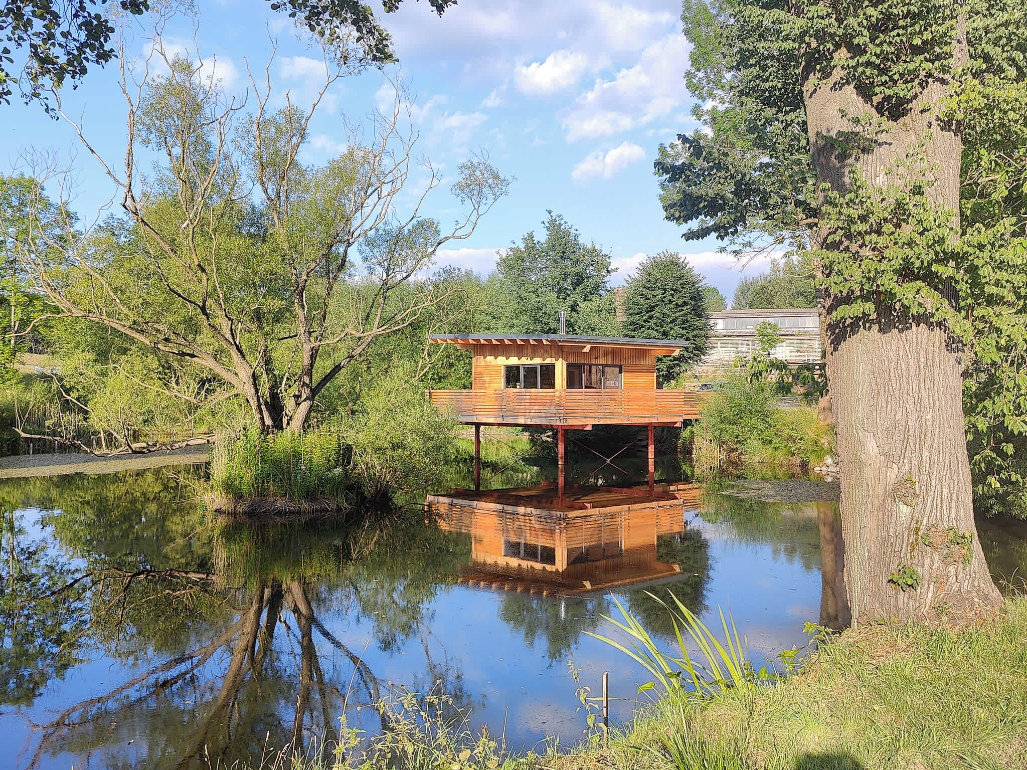 A wooden cabin with a deck, situated by a calm lake, surrounded by trees.