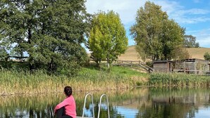A person sitting on a dock by a calm body of water, surrounded by trees and a wooden structure in the background. - Designer apartment with sauna, hot tub and fireplace (Großharthau)