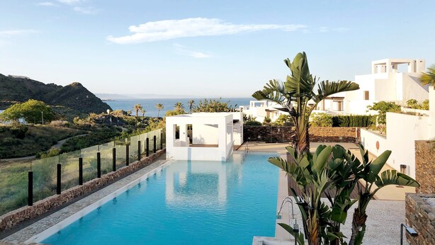 A swimming pool with a view of the sea, surrounded by white buildings and greenery.