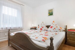 A double bed with floral bedding, a nightstand with a lamp, a radiator, and a framed picture on the wall.