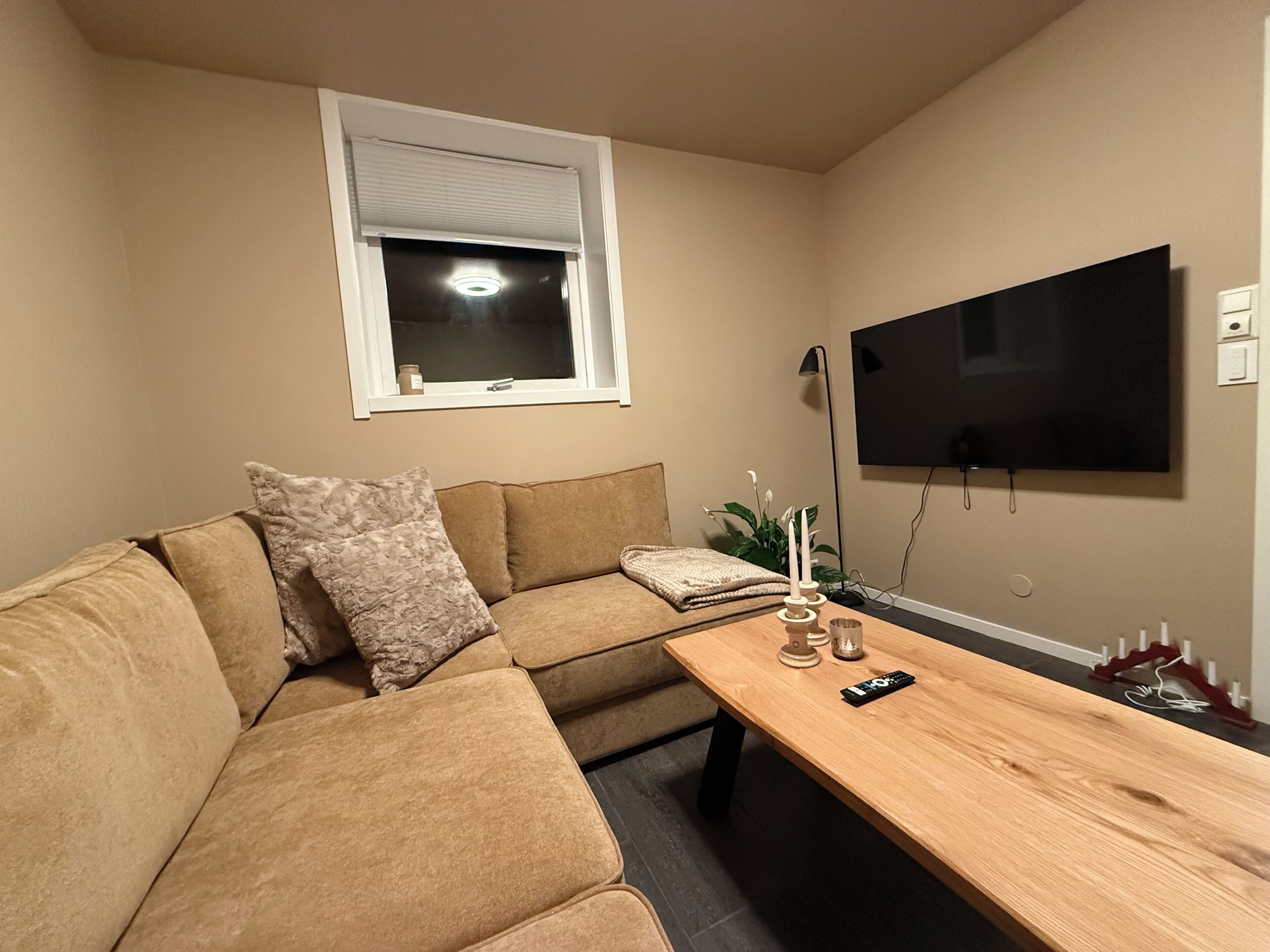 Een woonkamer met een hoekbank, een houten salontafel, een aan de muur bevestigde televisie en een raam met zonwering.