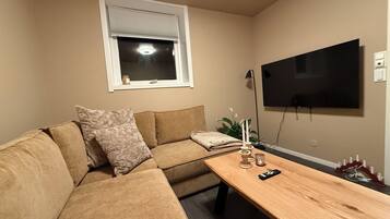 Een woonkamer met een hoekbank, een houten salontafel, een aan de muur bevestigde televisie en een raam met zonwering.