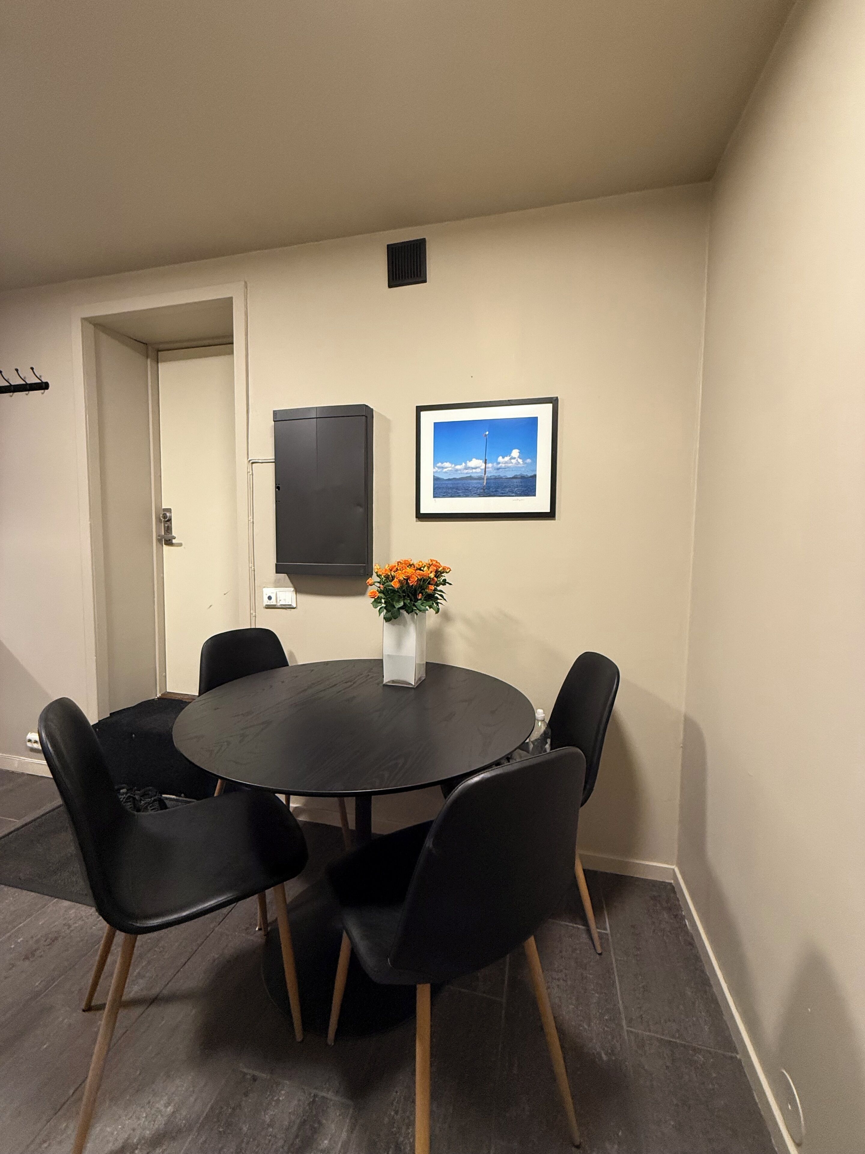 Une salle de conférence avec une table ronde, des chaises noires, un vase avec des fleurs oranges, un tableau encadré au mur et une porte donnant sur une autre pièce.