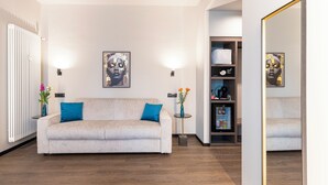 A modern living room with a beige sofa, two blue cushions, a small side table with a vase of flowers, a framed artwork on the wall, and a bookshelf with various items. - AH - Collezione Brera (Milan)