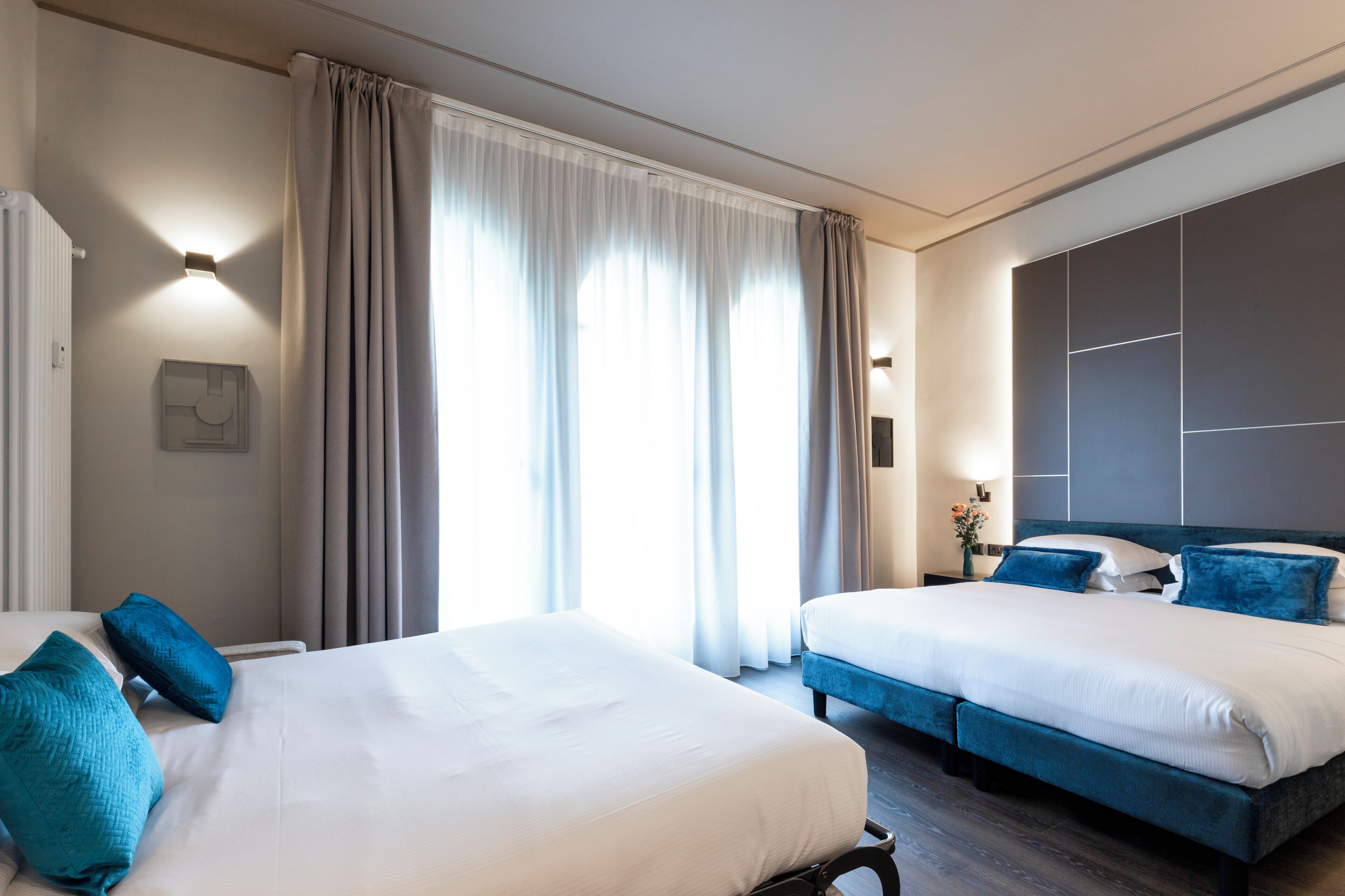 A hotel room with two beds, a large window with curtains, a wall-mounted TV, and a bedside table with a vase of flowers.