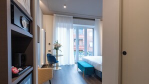 A hotel room with a bed, a desk with a chair, a small table with a plant, and a view of the outside balcony. - AH - Collezione Brera (Milan)