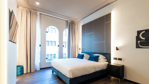 A modern bedroom with a large bed, a nightstand, and a view of the city through large windows. - AH - Collezione Brera (Milan)