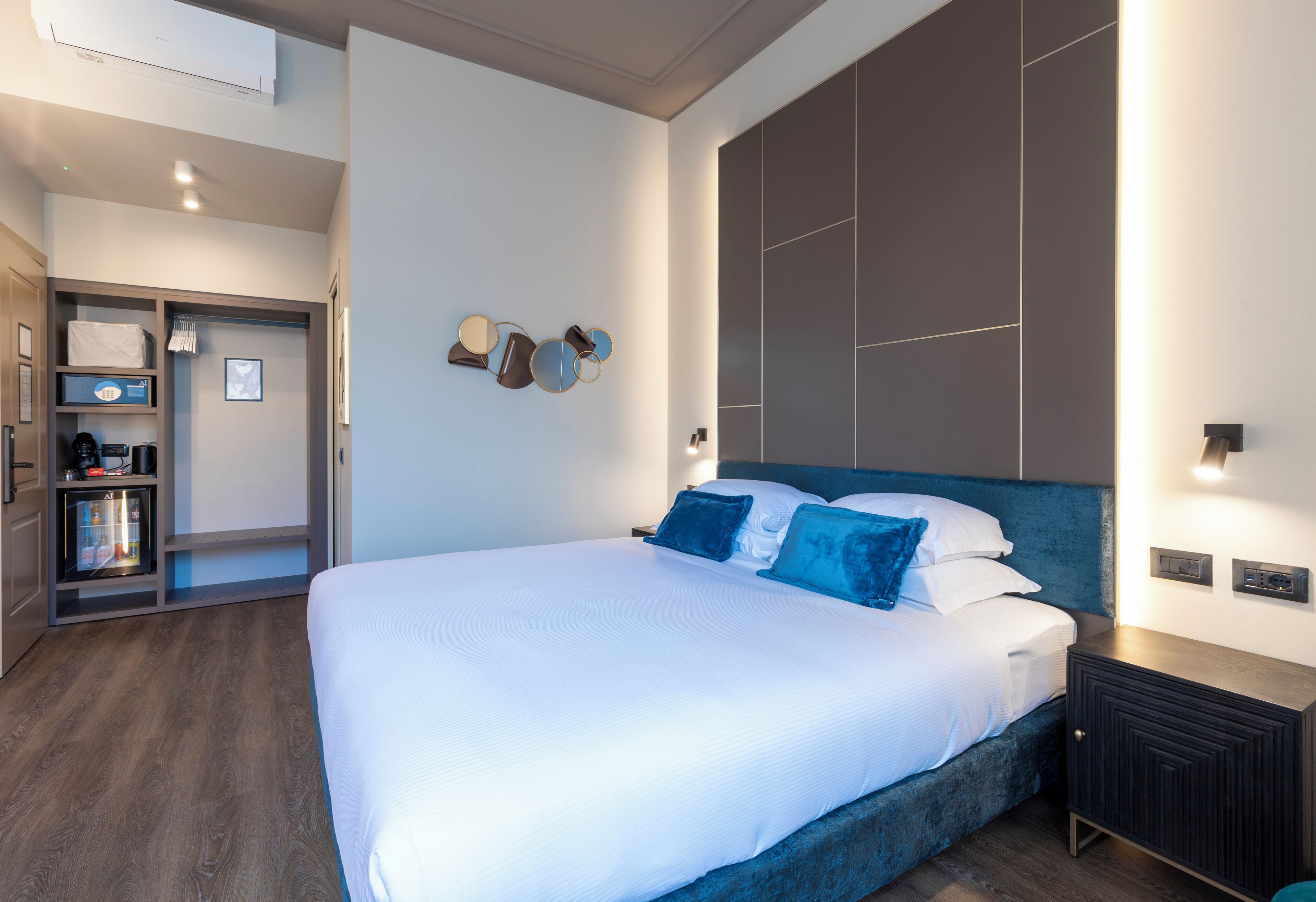 A modern hotel room with a large bed, a built-in wardrobe, a minibar, and a wall-mounted light fixture.