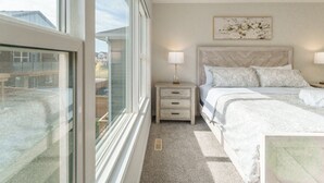 Une chambre avec une grande fenêtre, un lit recouvert d’une literie à motifs floraux, une table de chevet avec une lampe et une œuvre d’art accrochée au mur.