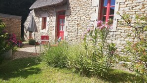 Ein Steinhäuschen mit roten Türen und Fenstern, umgeben von viel Grün und einem Garten.