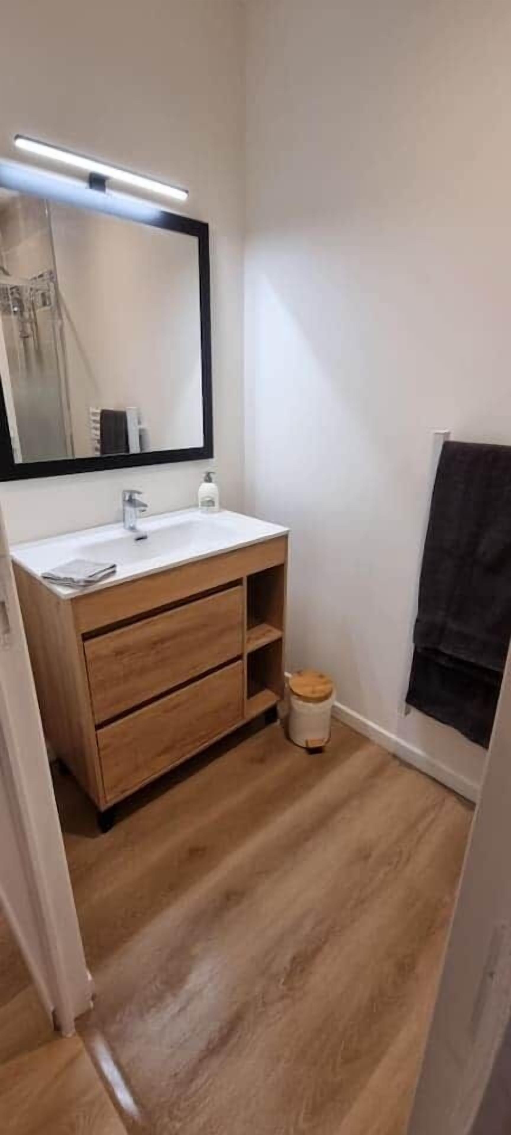 A small bathroom with a wooden vanity, a mirror, a white sink, and a black towel hanging on the wall.