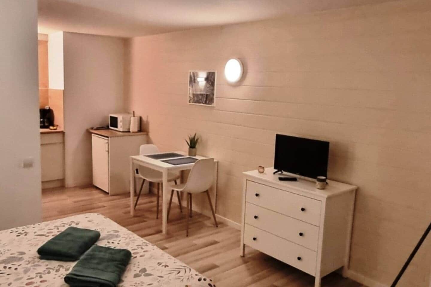 A compact room with a bed, a desk, a chair, a TV, and a small kitchenette.