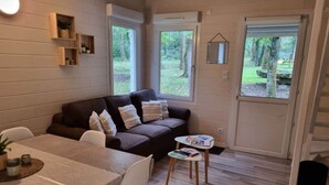 A cozy living space with a sofa, dining table, and chairs, a small coffee table, and a view of a garden through a sliding door. - Chalet 7 - 8P-Pool -100m Black Frame (Saumur)