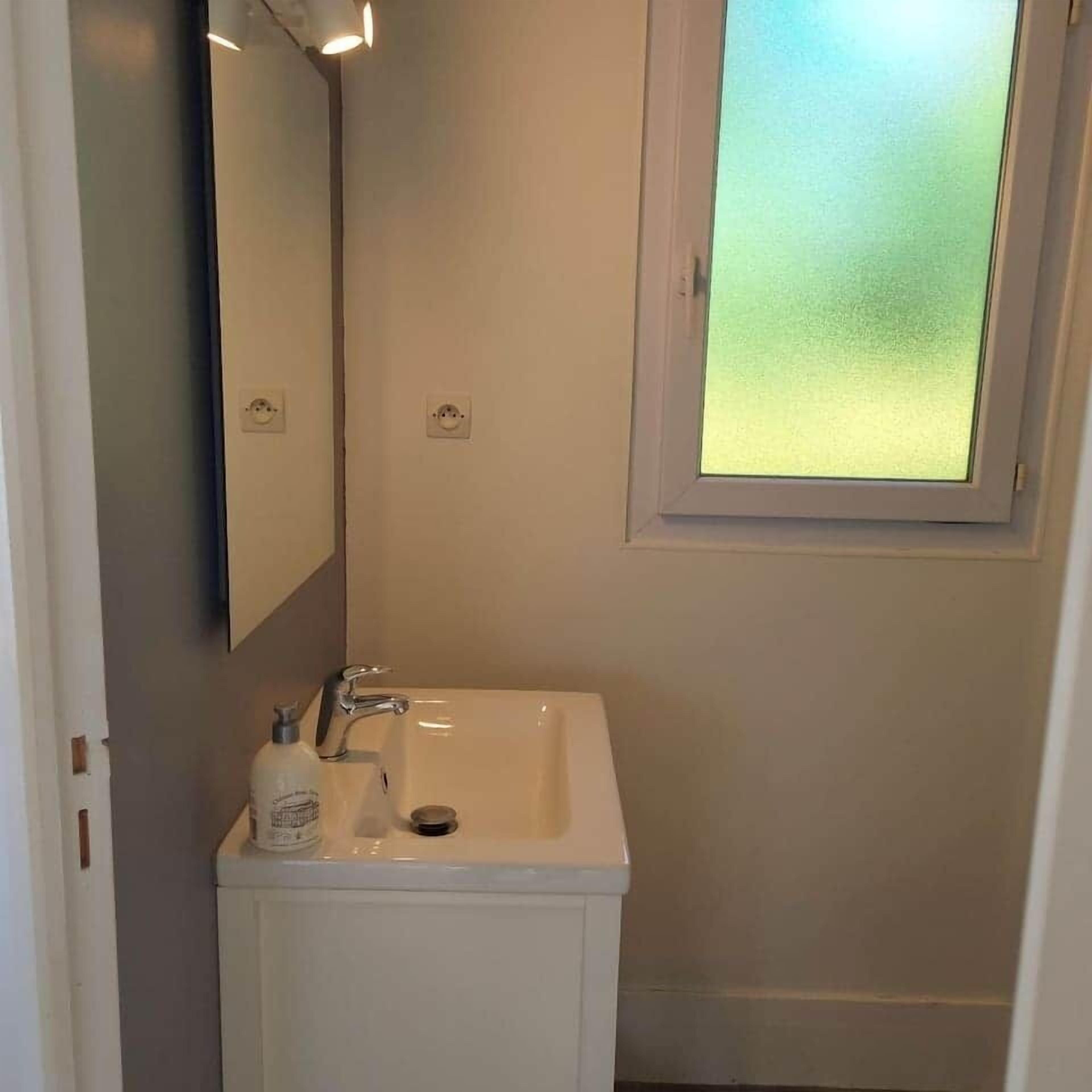 Une salle de bain avec un lavabo blanc, un miroir et une fenêtre en verre dépoli.