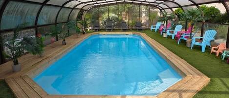 A covered swimming pool with a wooden deck, surrounded by grass and colorful chairs.