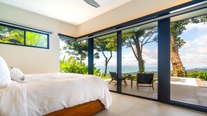 A bedroom with a large bed, a view of the outdoors through glass doors, and a seating area with a table and chairs. - Jungle Luxury Villa • 3BR w/ Pool & Ocean View (Savegre)