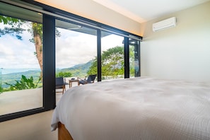 A bedroom with a large bed, a chair, and a table. It has a view of a mountainous landscape through large windows. - Jungle Luxury Villa • 3BR w/ Pool & Ocean View (Savegre)
