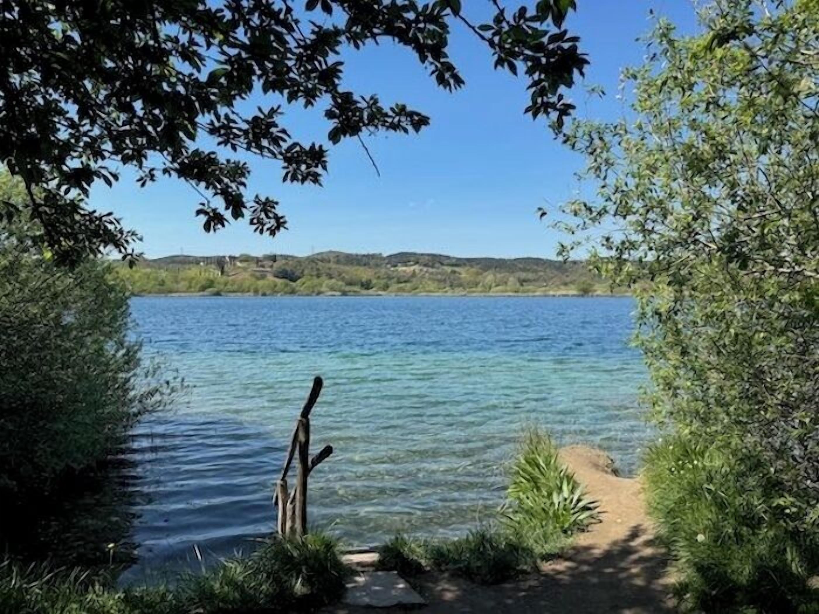Visão nítida de um lago com um caminho que leva até a margem, rodeado por vegetação e colinas ao fundo.