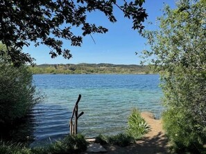 A clear view of a lake with a pathway leading to the water's edge, surrounded by greenery and hills in the background. - Alma del Lago Charming Apartments (La Pesta)