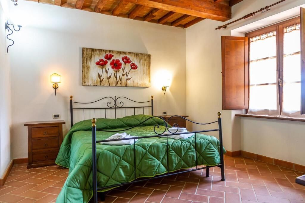 A bedroom with a green bed, wooden headboard, two bedside tables, a painting of red flowers, and a window with wooden shutters.