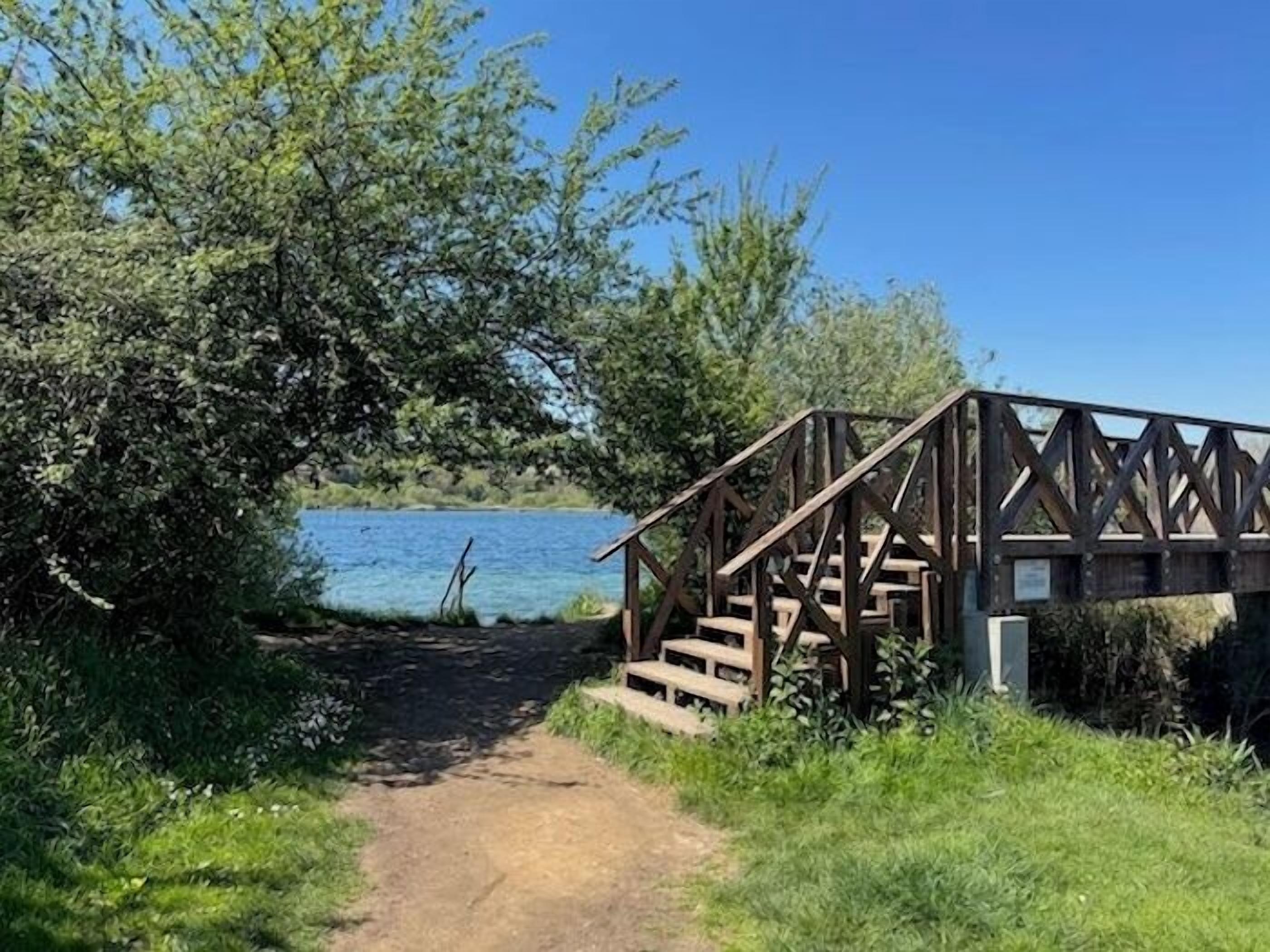 Eine Holztreppe führt zu einer Brücke über ein Gewässer, umgeben von viel Grün.