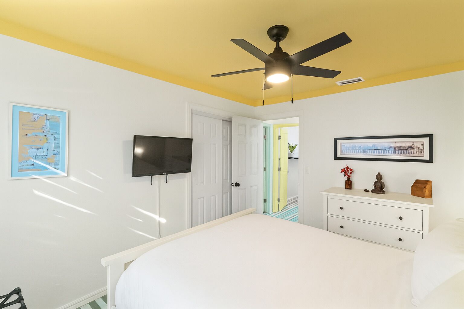 A bedroom with a bed, a ceiling fan, a TV, a dresser, and a door leading to another room.