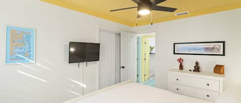 A bedroom with a bed, a ceiling fan, a TV, a dresser, and a door leading to another room.