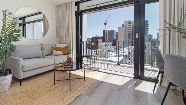A modern living room with a sofa, a coffee table, and a large window with a view of the cityscape and construction cranes. - The York | Light + Spacious CBD Apartment (Cape Town)