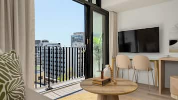 A modern living room with a large window, a flat-screen TV, a round wooden table, and a sofa with a patterned cushion.