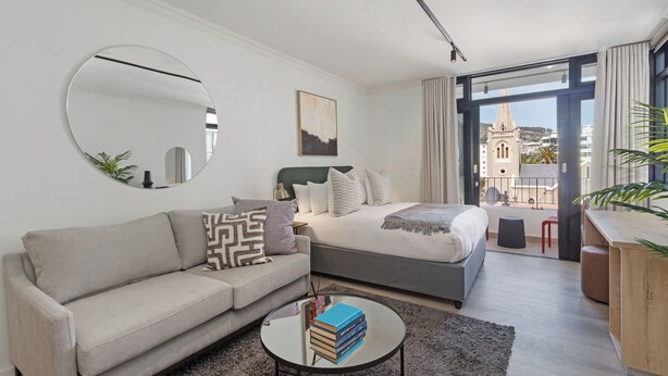 Een moderne hotelkamer met een bed, een slaapbank, een salontafel met boeken, een grote spiegel en uitzicht op een gebouw door het raam.