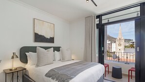 Een moderne slaapkamer met een groot bed, een nachtkastje met een lamp en een uitzicht op een stadsgezicht door een balkondeur.
