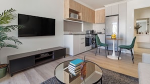 Een moderne woonkamer met een flatscreen televisie, een glazen salontafel met boeken, een potplant en een eethoek met twee stoelen en een kleine tafel.