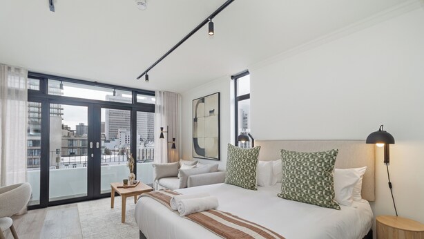 Habitación de hotel moderna con una cama grande, zona de estar con una mesa y vistas de la ciudad a través de grandes ventanales.