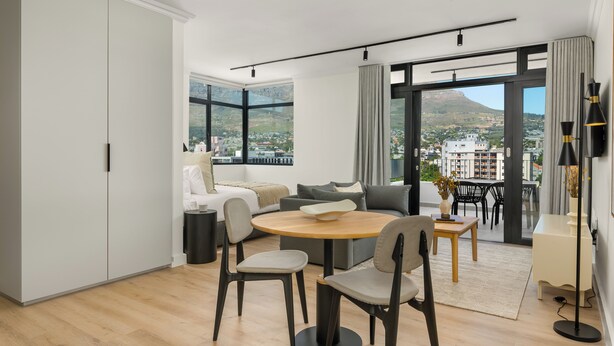 A modern living room with a round wooden table, two grey chairs, a grey sofa, and a large window offering a city view. - The York | Spacious CBD Apartment (Cape Town)
