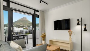 A modern living room with a sofa, a coffee table, a TV mounted on the wall, and a view of a cityscape and a mountain. - The York | Spacious CBD Apartment (Cape Town)