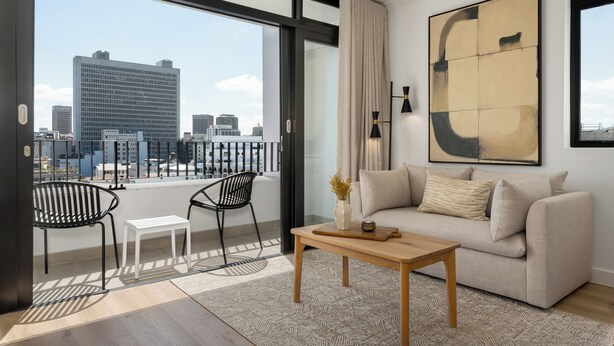 A modern living room with a sofa, a coffee table, and two chairs. A large abstract painting is on the wall, and there's a view of the city through a glass door.