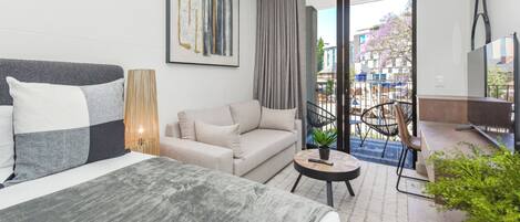Una habitación de hotel moderna con una cama, sofá y una mesita con una planta. Hay un ventanal con vistas al exterior.